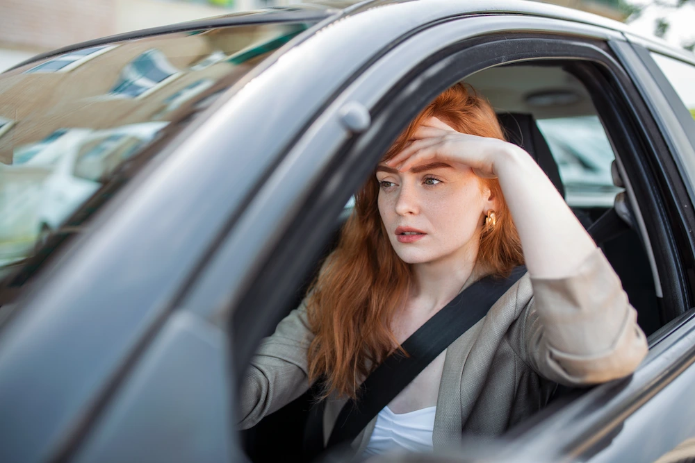 A woman with red hair and freckles sits in a car, hand on her forehead, looking out the window with a worried expression.