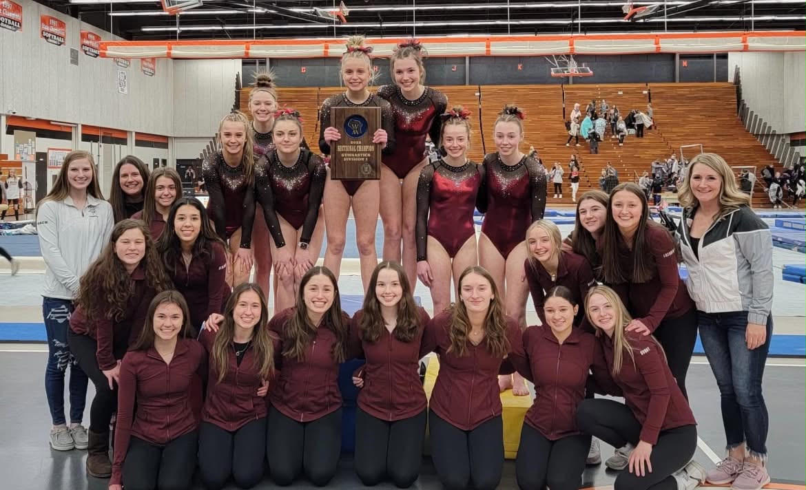 A group of approximately 20 female gymnasts and coaches smiling and posing with a "2023 Sectional Champion" trophy in a gymnasium.