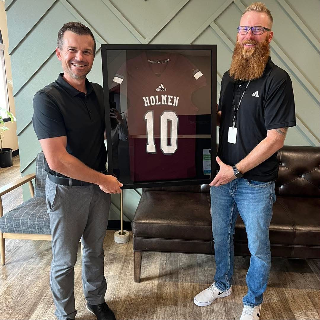Two smiling men holding a framed maroon sports jersey with 'HOLMEN 10' in white text.