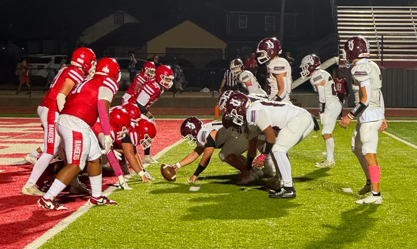 An American football game at night, showing two teams in red and maroon uniforms lined up at the line of scrimmage.
