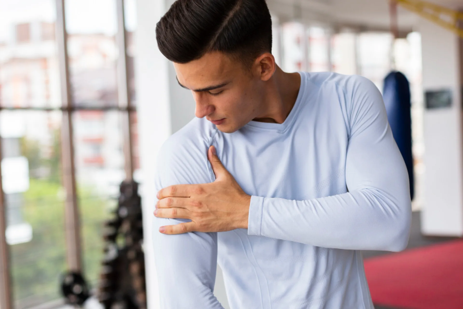 A man in a light blue long-sleeved shirt holds his left shoulder with his right hand, looking down at it in a gym.